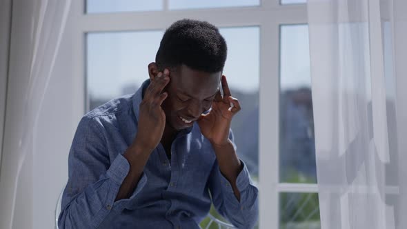 Overwhelmed Young Man Rubbing Temples Sitting in Sunny Home Office Indoors