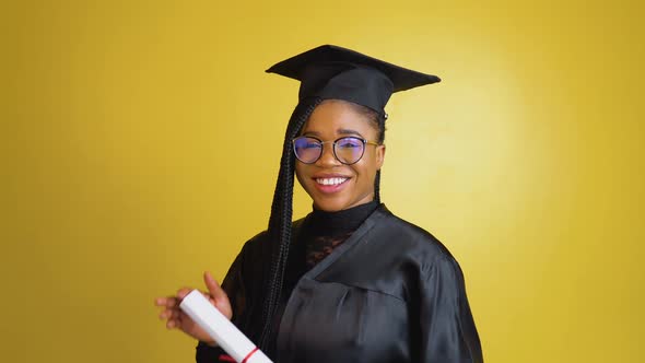 The African American Graduate Student in Magister's Robe Stands on Yellow Background and Holds in