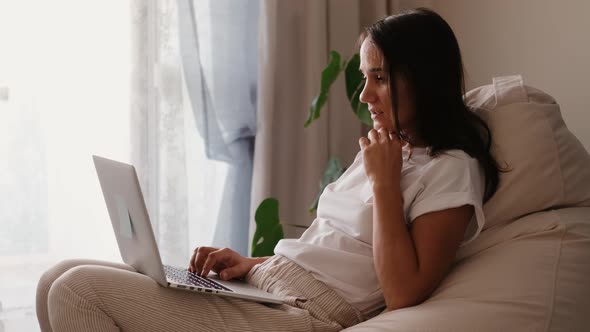 Excited Young Winner Woman Looking at Laptop Celebrating Internet Success Sitting on a Pouf at Home