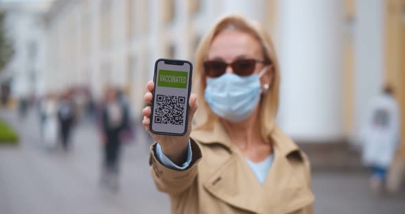 Mature Woman in Protective Medical Mask Holding Cellphone with Electronic Passport of Vaccination