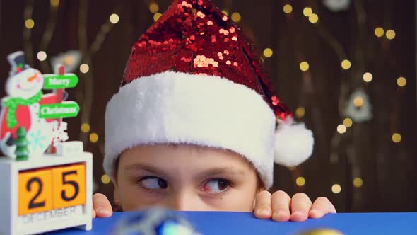 Smiling little girl in Santa hat looking out from behind table. Child at Christmas time