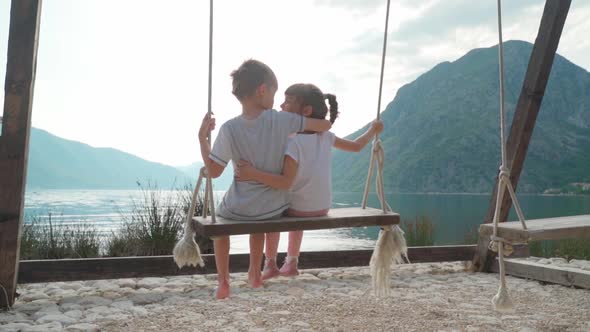 A Girl and a Boy Swing on a Swing Near the Sea with a Beautiful View of the Bay and Mountains