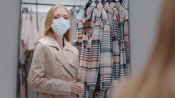 Young Millennial Woman in Protective Medical Mask Chooses New Outfit in Store Trying Autumn Coat