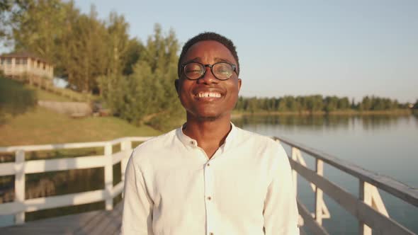 A Young Black Guy in Glasses Stands on a Wooden Pier on the River Bank and Closing His Eyes Enjoys