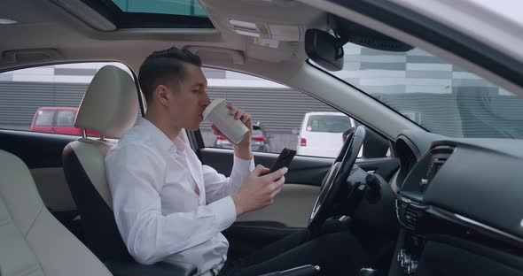 Portrait of Young Man Entrepreneur Sits in the Driver's Seat in a Car and Uses Smartphone for