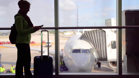 Silhouette of woman smiling with luggage and smartphone with blurry plane through window outside.