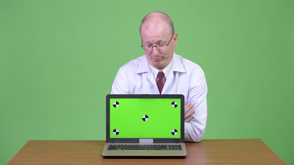 Upset Mature Bald Man Doctor Showing Laptop Against Wooden Table