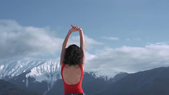 Young Curly Woman in Snowboard Goggles Standing Back and Raising Hands Up at Winter Ski Resort at