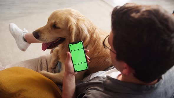 Brunette Man Wearing Glasses Sitting on Sofa with Golden Retriever Using Smartphone for Buying