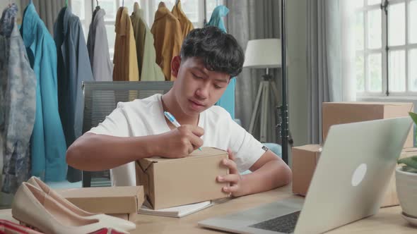 Asian Man Online Seller Writing On Package While Using Computer For Selling Clothes At Home