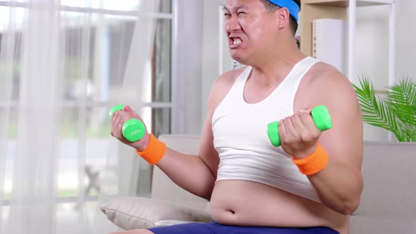 Funny fat Asian guy in sportswear lifting two side dumbbell with difficultly in living room