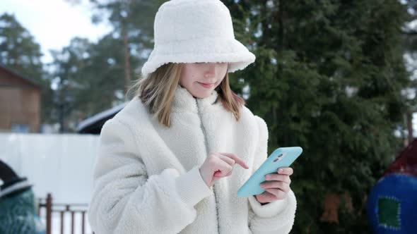 Beautiful Young Girl in White Furry Jacket and Hat Using Smartphone Outdoor