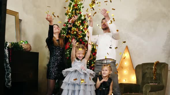 Young Happy Family of Four Standing By the Christmas Tree and Throwing Up Golden Confetti