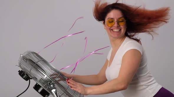 Redhaired Caucasian Woman in Sunglasses Enjoys the Breeze By the Electric Fan on a White Background