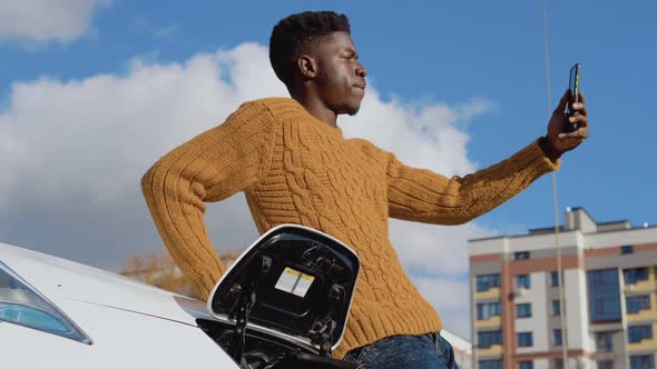 A Black Driver is Photographed on a Smartphone Camera Standing Near a White Electric Car