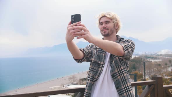 Young Stylish Blonde Man Takes Selfie on the Background of the Sea