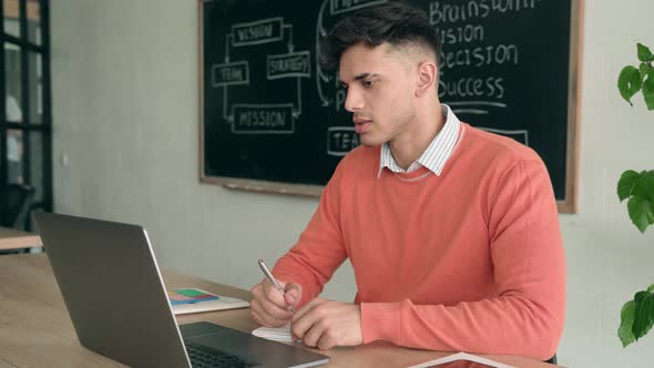 Serious Indian Student Has Video Chat at Pc at Desk in Classroom Writing Notes