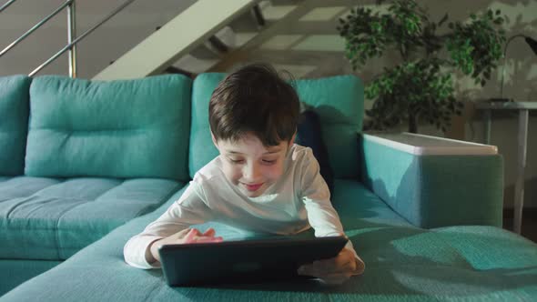 Curious Cute Elementary School Kid Boy Using Digital Tablet Technology Device Lying On Sofa Alone