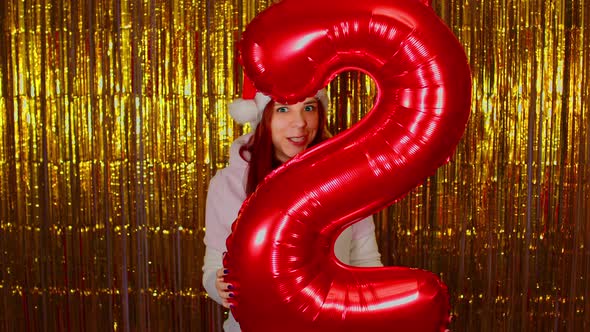 Woman in Santa Hat Holds Red Figure Two on Background of Shimmering Golden Tinsel