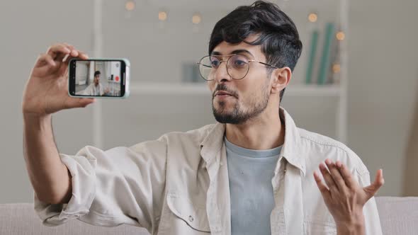 Young Cheerful Arabic Guy Bearded Man Wearing Glasses Sitting at Home Indoors Couch Using Mobile