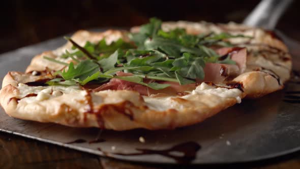 Camera follows putting a white pizza with prosciutto and arugula on a table. Slow Motion.