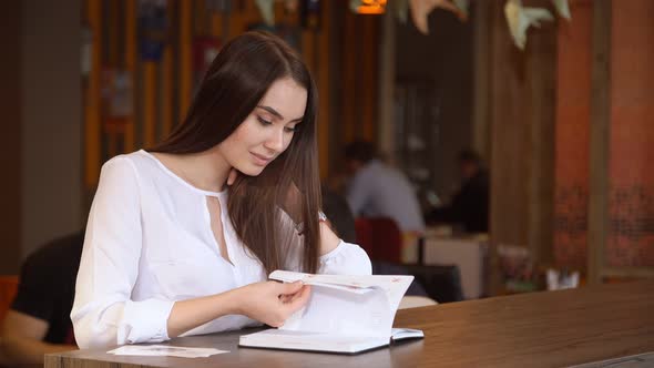 Young Businesswoman in Cafe Leafing Through the Notebook.