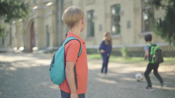 Camera Approaches To Cute Caucasian Boy Looking Back at Classmates Playing Soccer and Showing Thumb