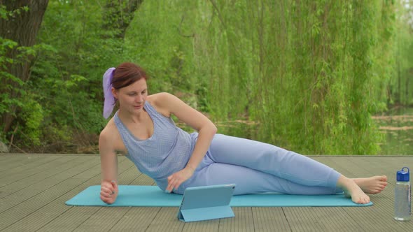 30s Woman in Sportswear Doing Aerobics Plank Exercise Using Tablet Computer Outdoors