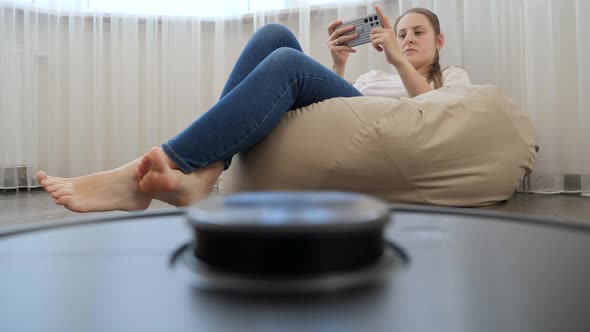 POV Shot of Robot Vacuum Cleaner Doing House Cleanup While Young Woman Relaxing and Using Smartphone