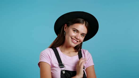 Attractive Hipster Playful Woman in Overall Wear and Hat Smiling, Flirting To Camera Over Blue Wall