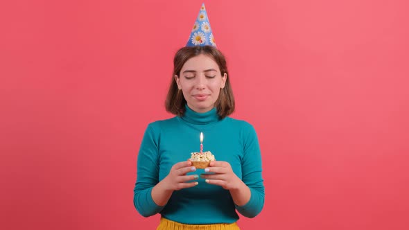 Young Woman in Birthday Hat Blowing Out Candle on Cake, Isolated on Red Background.