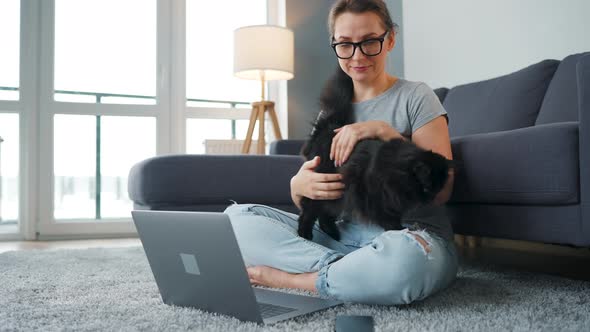 Casually Dressed Woman Sits on a Carpet with a Laptop Holds on Her Knees and Strokes a Fluffy Cat