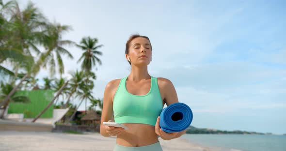 Woman Stands on the Beach with Fitness Mat and Taking a Dip Breath She Listening Favourite Music