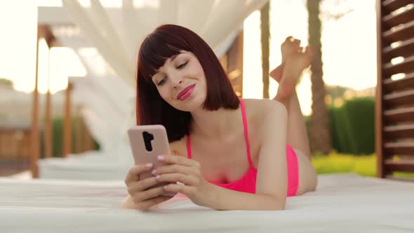 Joyful Young Lady in Pink Swimwear Lying in Gazebo Near the Resort Pool and Using Smartphone