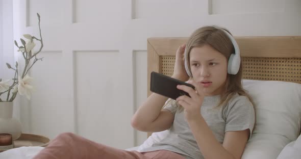 School Girl Is Lying on Bed in Room and Playing Game on Smartphone By Internet, Listening To Music