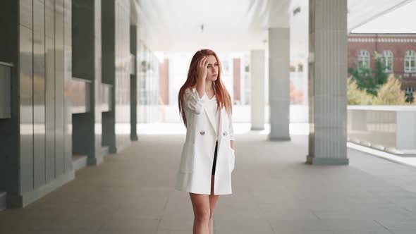 Beautiful business woman red hair in white coat stands near an office building