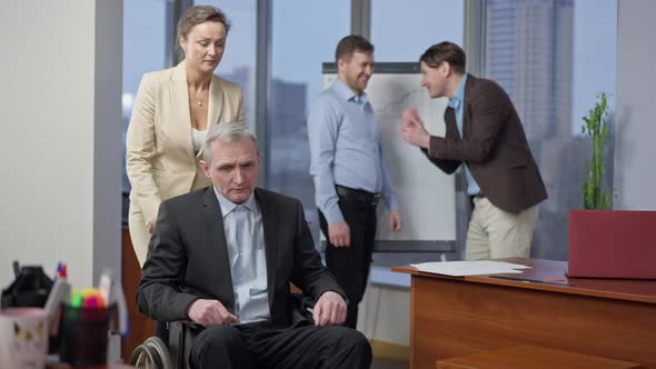 Young Beautiful Woman Rolling Wheelchair with Disabled Senior Man in Office As Male Workers Talking