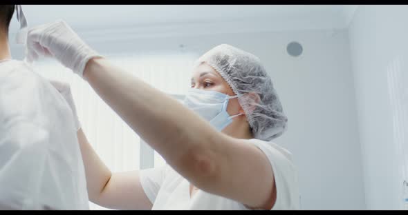 A Nurse Helps a Dentist Put on a White Sterile Suit Tying the Ties of His Back