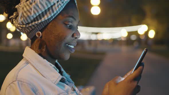 Smiling African Woman Typing on Smartphone Outdoors in Evening