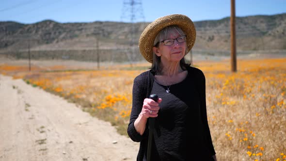 An aging woman smiling and walking down a dirt road in the summer heat with orange flowers in the fi