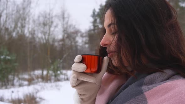 Young Cute Swarthy Brunette Woman Drinking Hot Drink From Orange Thermos Cup in Snowy Winter Forest