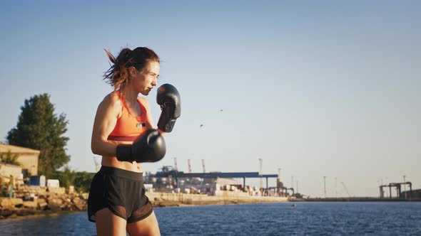 Young Female in Boxing Gloves and Sportswear is Boxing While Training By Seaside Near the City Port