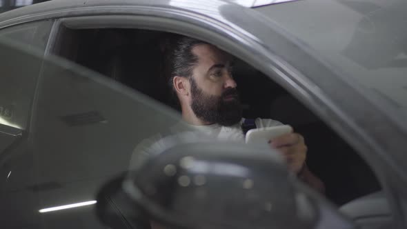 Professional Mechanic Sitting Inside the Car and Verifies Information on His Phone. Bearded Man in