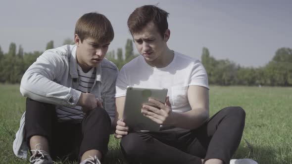 Two Concentrated Caucasian Friends Sitting on Green Summer Lawn Using Tablet