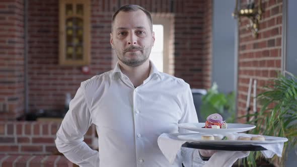 Portrait of Male Waiter Looking at Camera Walking in Slow Motion Holding Tray with Dessert and Salad