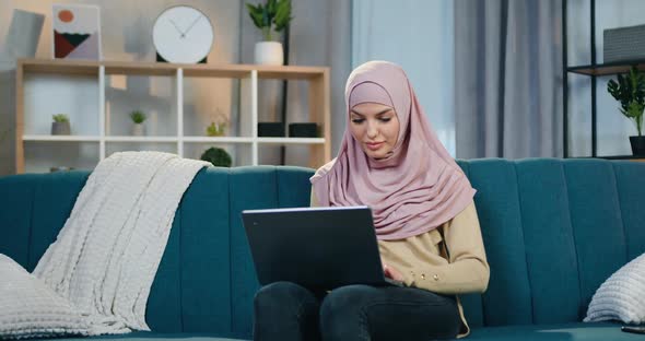 Arabian Woman in Hijab Sitting on Soft Couch in Contemporary Apartment and Uses Computer