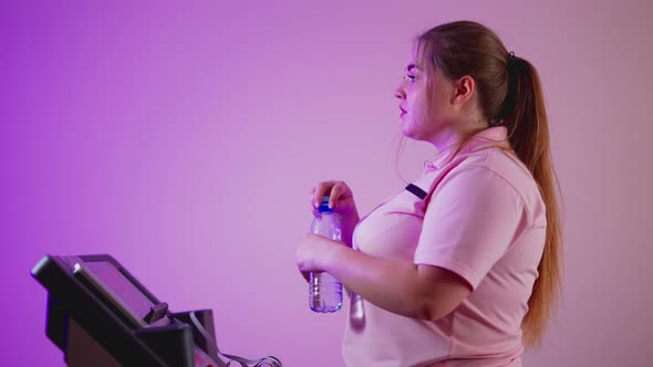 Side View of Perspiring Overweight Young Woman Walking on Treadmill Drinking Refreshing Water
