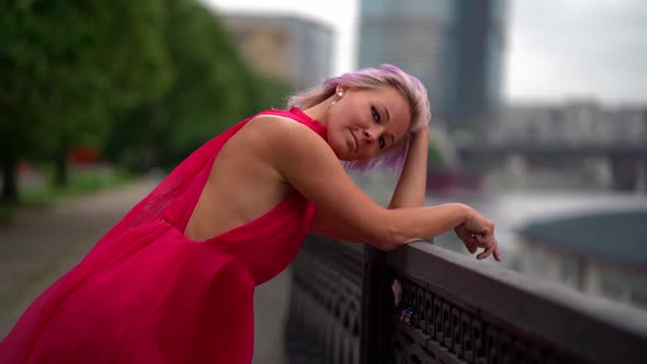 Blonde Woman Is Leaning on Fence of City Embankment and Looking at Camera