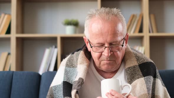 A Grayhaired Grandfather is Sitting on the Sofa Covered with a Gray Blanket and Drinking Warm Tea