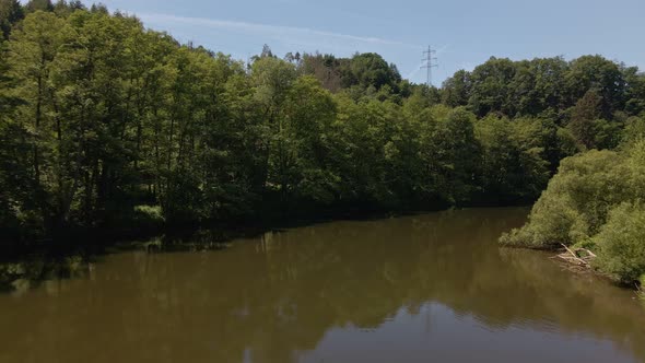 Wide angle drone footage following a meandering brown river in between lush green banks covered by t